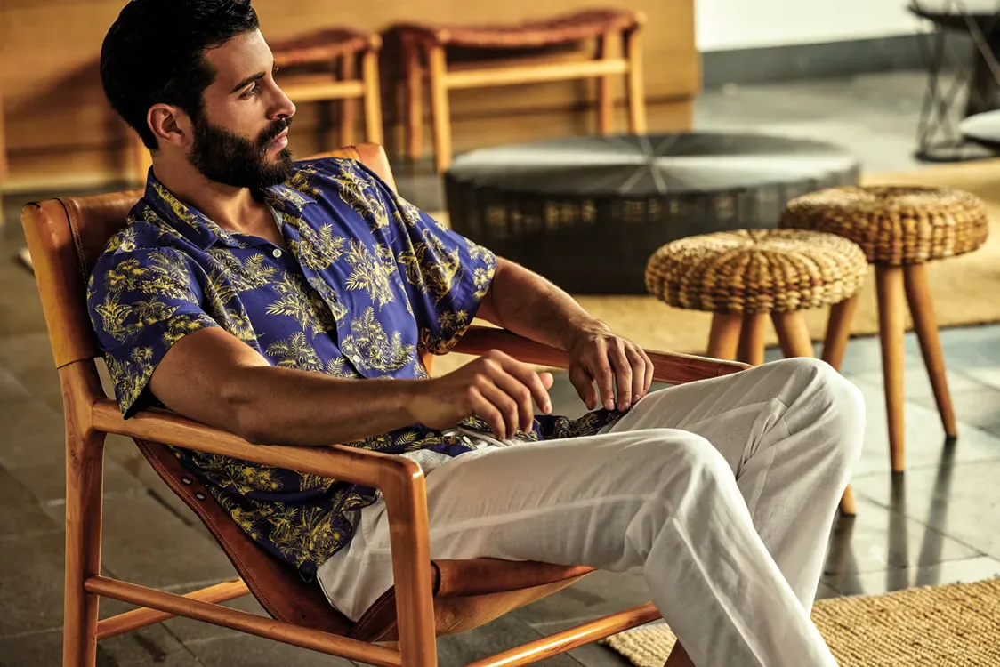 A man in a David printed shirt and linen trousers in a leather chair